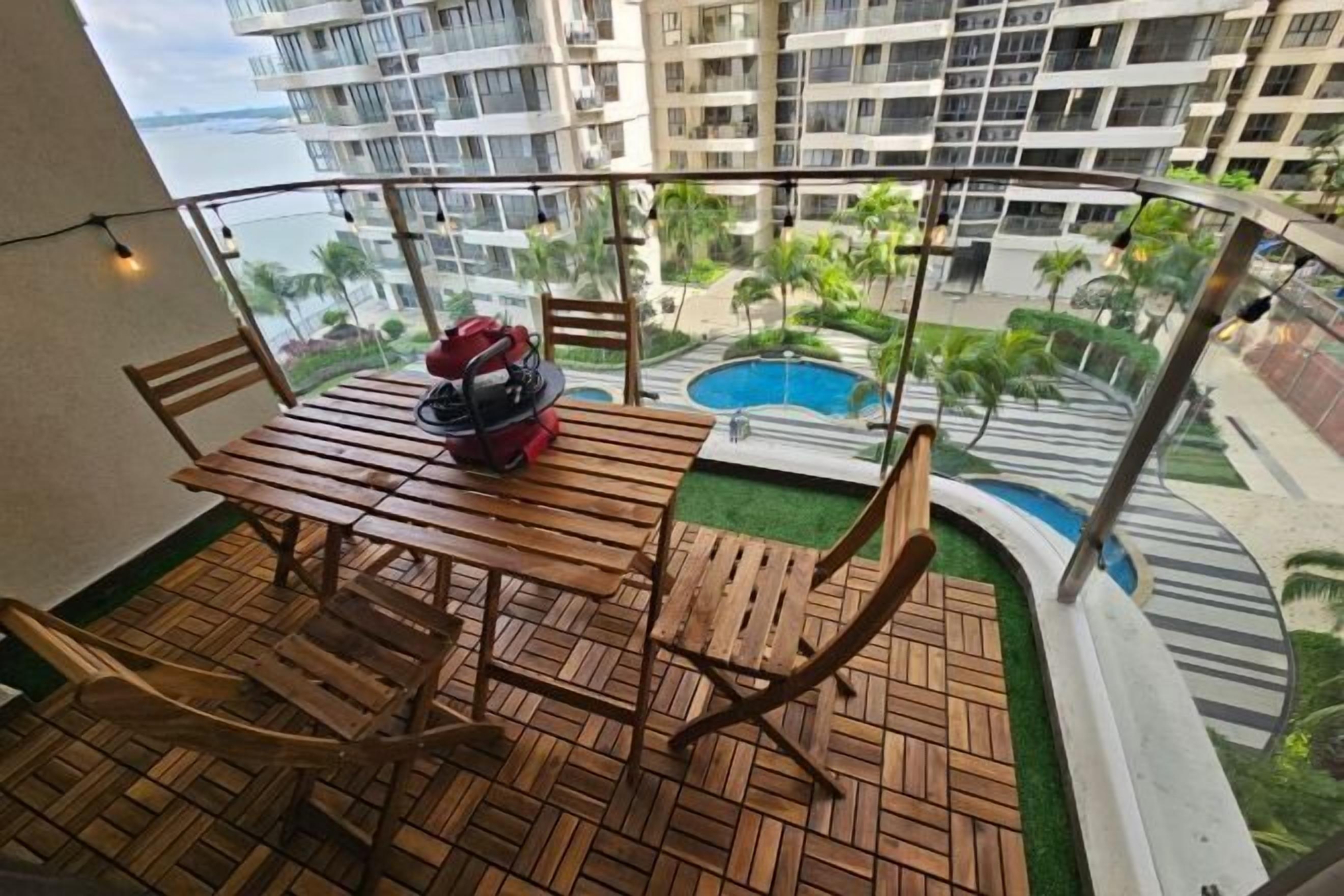 Ein Balkon mit Holztisch und Stühlen, einer roten Tasche und Blick auf ein Schwimmbecken und Gebäude.