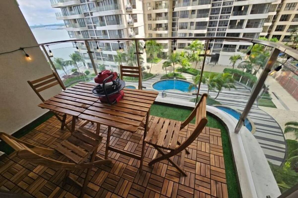 Uma varanda com mesa e cadeiras de madeira, uma bolsa vermelha e vista para uma piscina e edifícios.