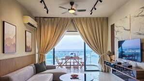 Una sala de estar moderna con un sofá, una mesa de centro redonda y un televisor. Hay un balcón con una mesa y sillas, y vistas al mar.