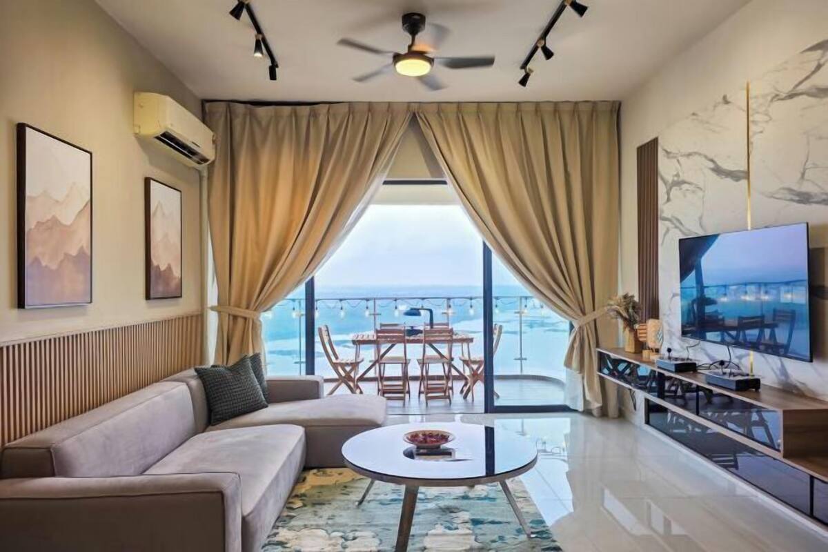 Uma sala de estar moderna com um sofá, uma mesa de centro redonda e uma televisão. Há uma varanda com uma mesa e cadeiras, e vista para o mar.