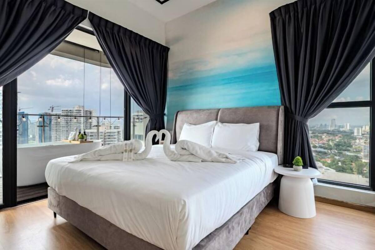 Quarto de hotel moderno com uma cama grande, cortinas pretas, vista da cidade e uma pequena mesa lateral com uma planta.