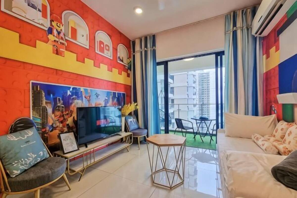 A living room with a TV, sofa, and chairs, a colorful mural on the wall, and a balcony with a view.