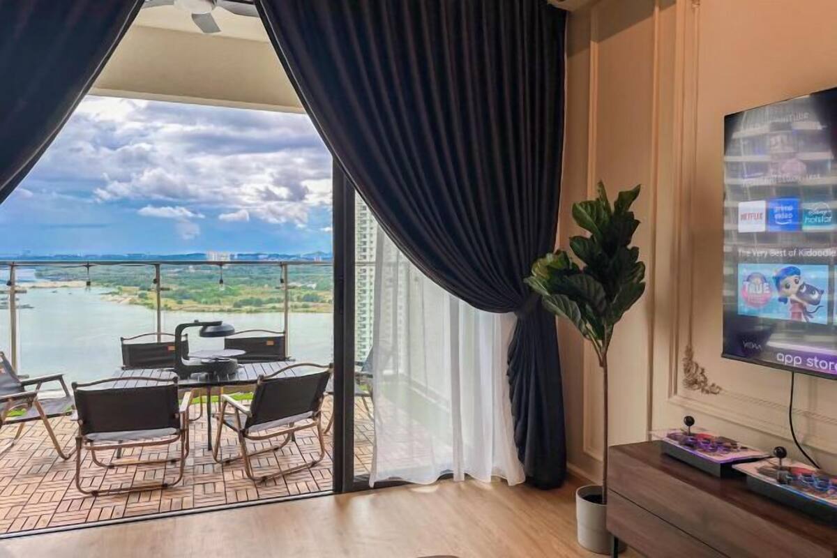 Quarto com varanda vista para um corpo d'água, área de jantar com cadeiras e mesa, e uma TV na parede.