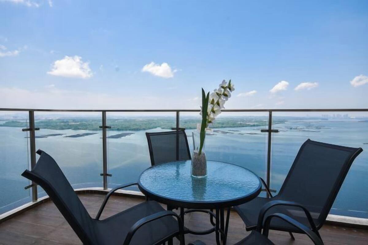 Uma varanda com mesa e cadeiras de vidro, um vaso com planta, e vista para um corpo d'água e um horizonte urbano distante.