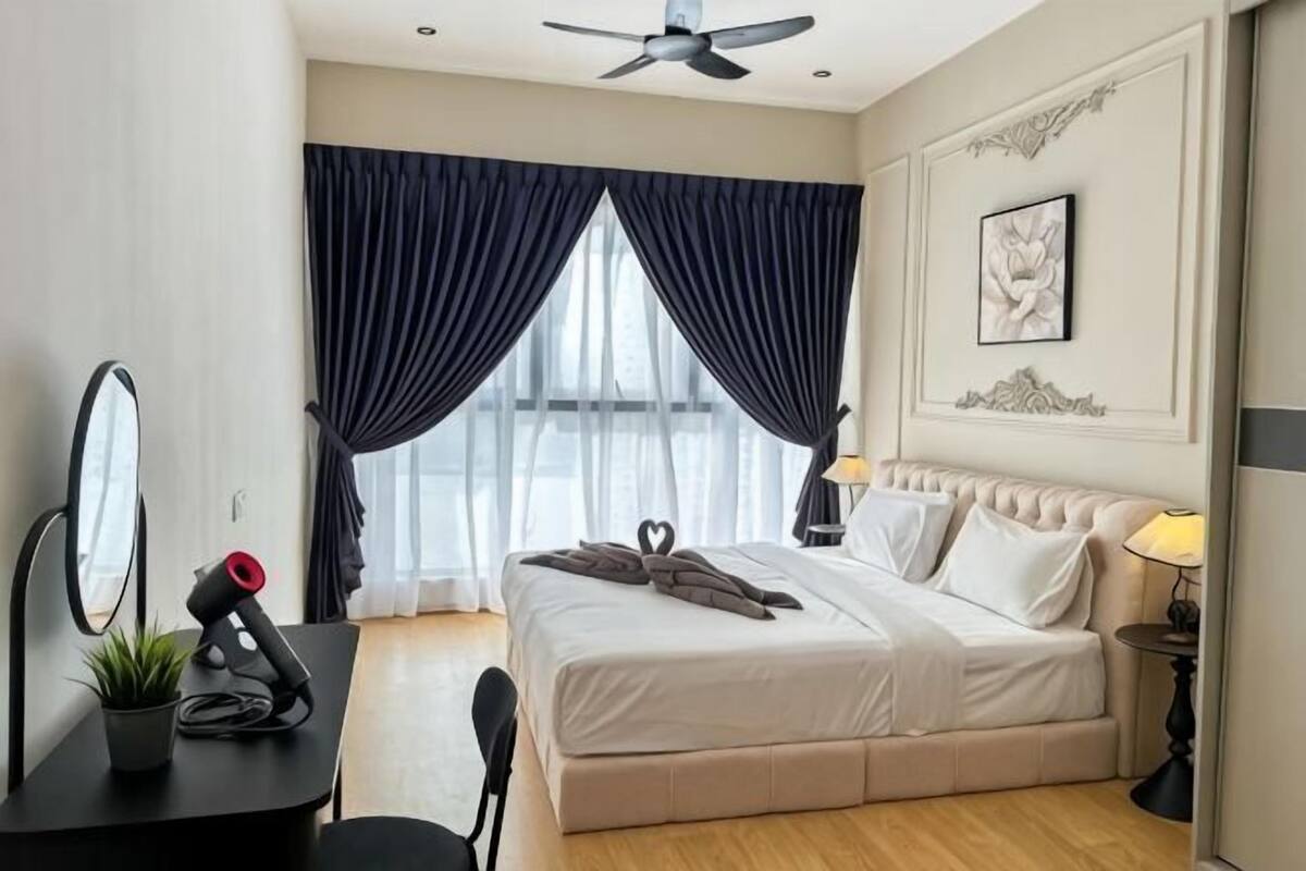 A hotel room with a bed, a desk with a mirror, a ceiling fan, and a window with curtains.
