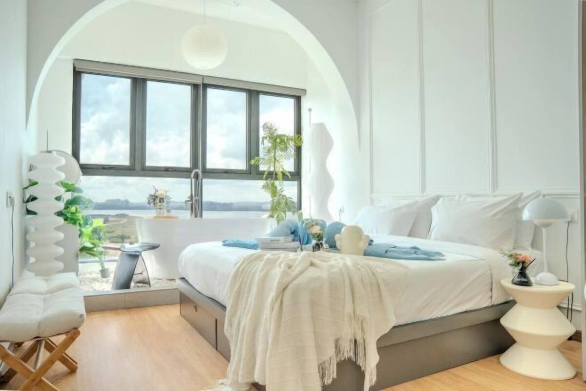 Quarto moderno com uma grande janela, uma cama, uma poltrona e uma mesinha de cabeceira.
