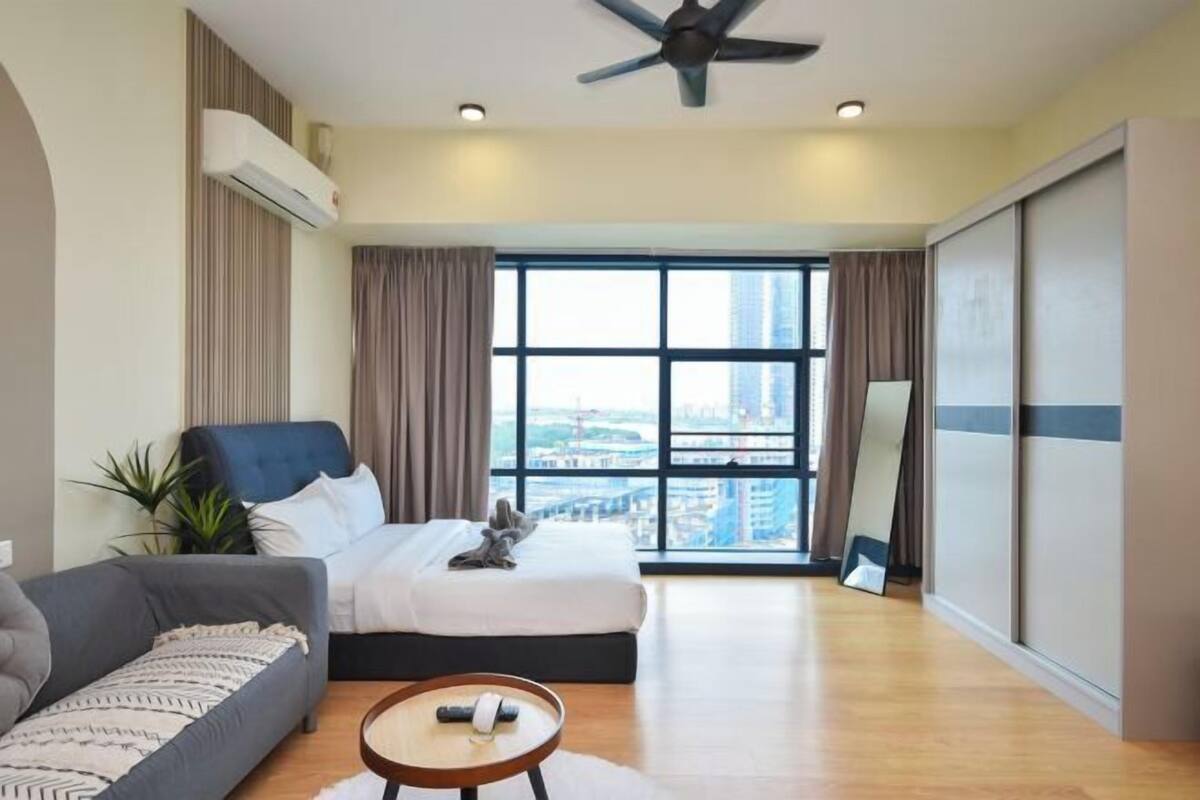 Quarto de hotel moderno com uma cama grande, um sofá, uma mesinha de centro e vista para a cidade através de amplas janelas.