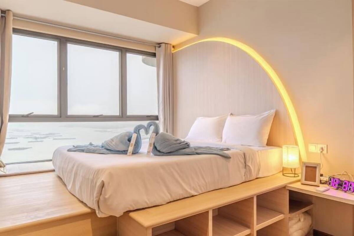 Quarto de hotel com uma cama, mesa de cabeceira com abajur e vista para o mar através de grandes janelas.