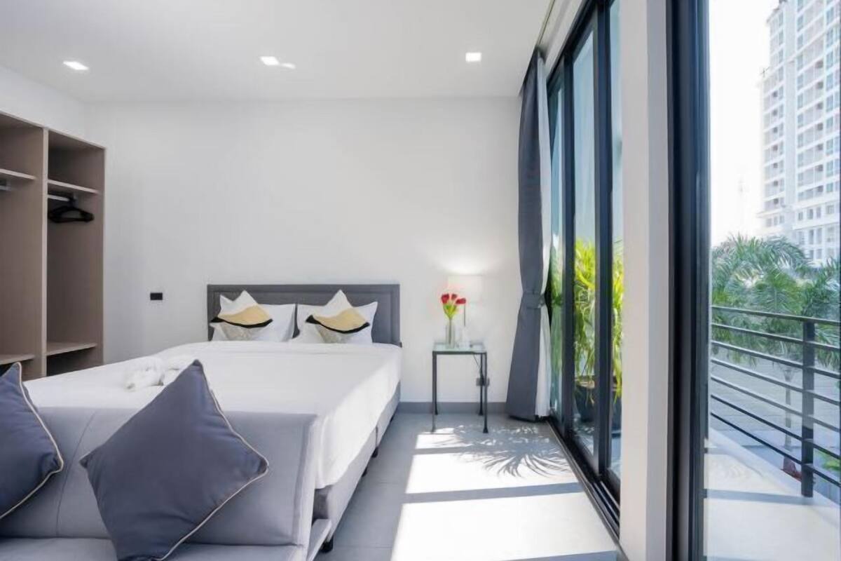 A modern hotel room with a large bed, a bedside table with a vase, and a view of a balcony with greenery.