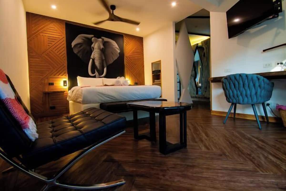 Ein modernes Hotelzimmer mit einem Bett, einer Sitzecke mit einem Sessel, einem kleinen Tisch, einem Deckenventilator und einem an der Wand befestigten Fernseher.