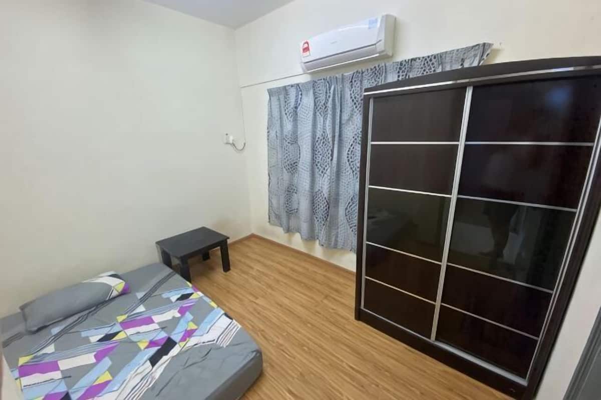 Ein Zimmer mit einem Bett, einem kleinen Tisch, einem Kleiderschrank, einem Fenster mit Vorhängen und einer Klimaanlage.