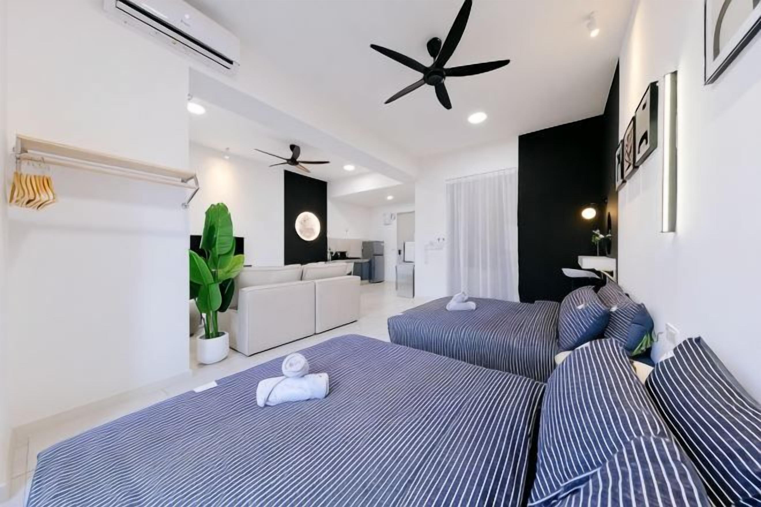 A modern hotel room with a bed, a ceiling fan, a plant, and a kitchenette in the background.