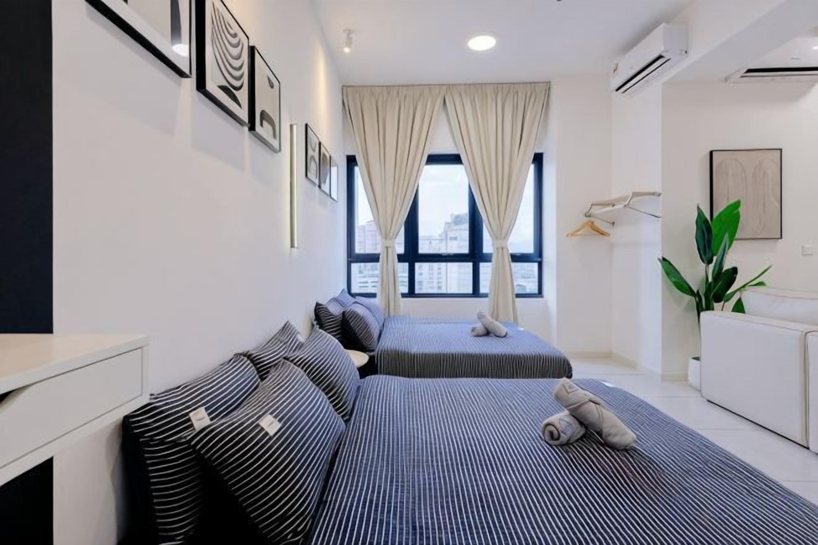 A hotel room with two beds, a desk, a chair, a plant, and a window with curtains.