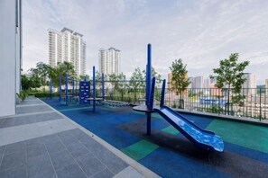 Un espace de jeux extérieur avec un toboggan et des structures d’escalade, entouré d’une clôture et d’arbres, avec des bâtiments en arrière-plan.