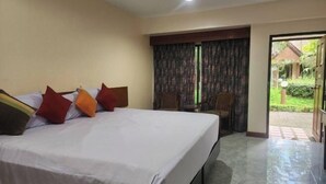 A hotel room with a large bed, a small table, and a view of the garden through a door. - Amorn inn resort (Chiang Rai)