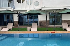 A swimming pool with lounge chairs and umbrellas, a building with a modern design, and a clear sky. - 1CE HOTEL (Puerto Galera)