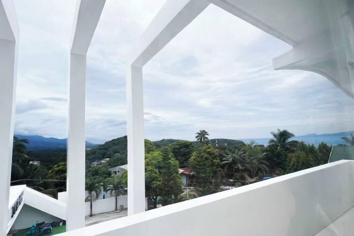 A modern building with white structural elements and a balcony overlooking a lush landscape with trees and distant hills.
