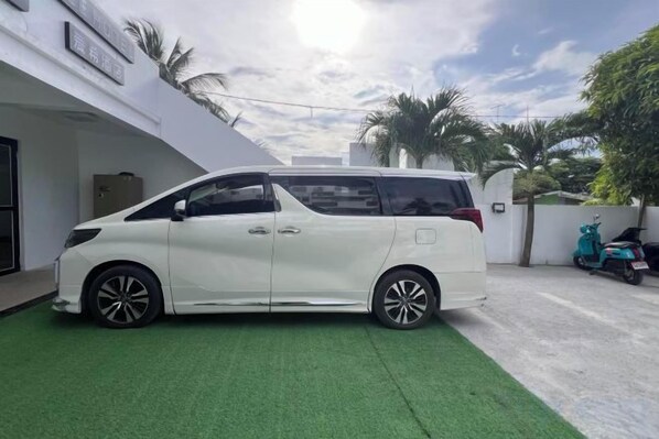 Una minivan blanca estacionada en una entrada con césped sintético verde, un edificio blanco con fachada curva, y un cielo despejado.