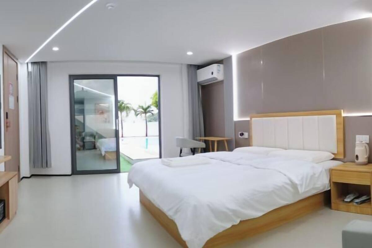 A modern hotel room with a large bed, a small table, a bedside table, and a sliding glass door leading to a balcony.