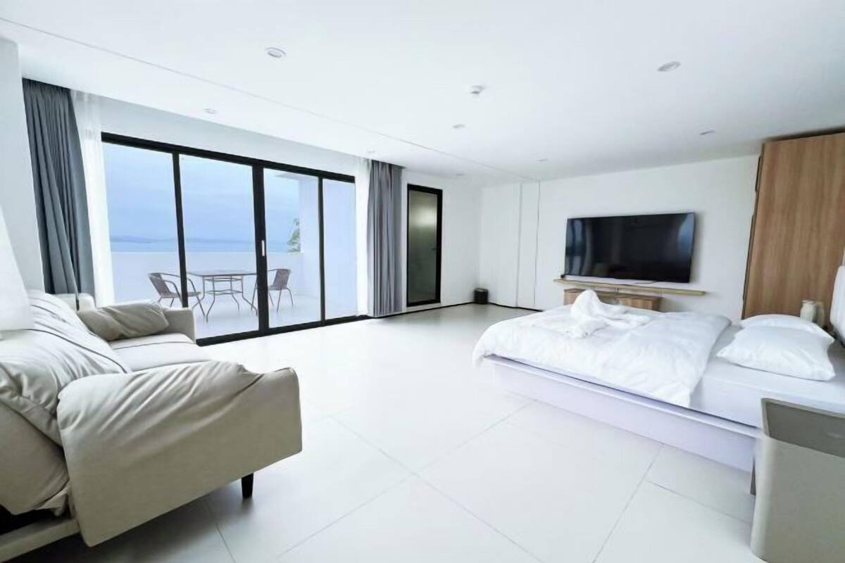 A modern hotel room with a large bed, a sofa, a TV, and a balcony with a view of the sea.