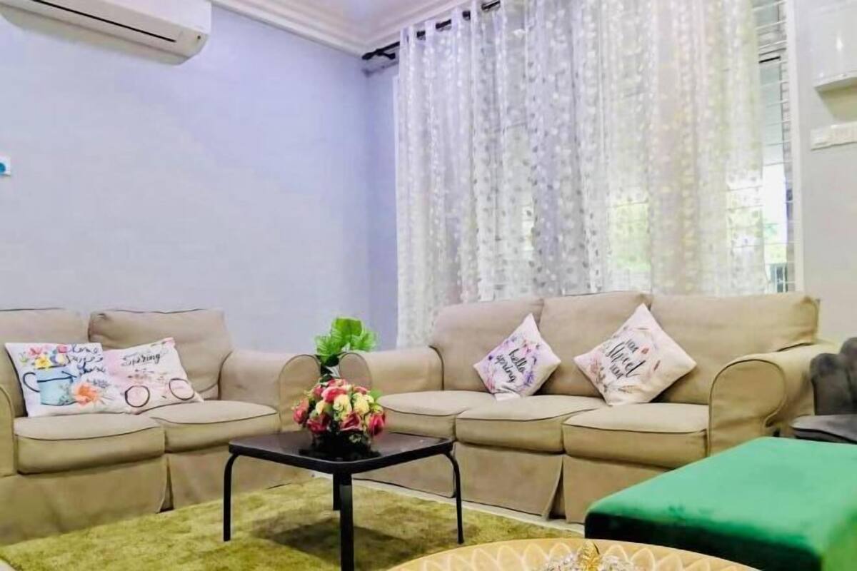 A living room with a beige sofa, a small table with a vase of flowers, and a green ottoman.