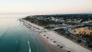 An aerial view of a coastal area with a beach, a road curving along the coast, and buildings in the background. - Dolce Vita, Esplanade (Aldinga Beach)