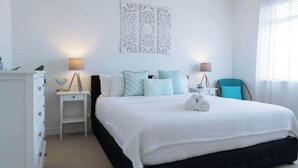 A neatly arranged bedroom with a large bed, two bedside tables, a chair, and a decorative wall piece. - Dolce Vita, Esplanade (Aldinga Beach)