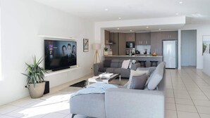 A modern living room with a grey sofa, a coffee table, and a TV on the wall. There is a kitchen in the background with grey cabinets and a refrigerator. - Dolce Vita, Esplanade (Aldinga Beach)