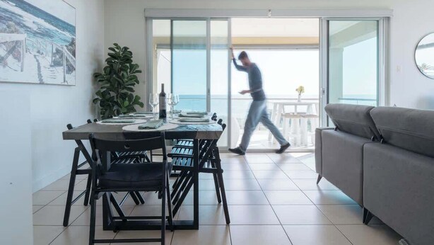 A modern living room with a grey sofa, a dining area with a table set for four, and a large sliding glass door leading to a balcony with a view of the sea. - Dolce Vita, Esplanade (Aldinga Beach)
