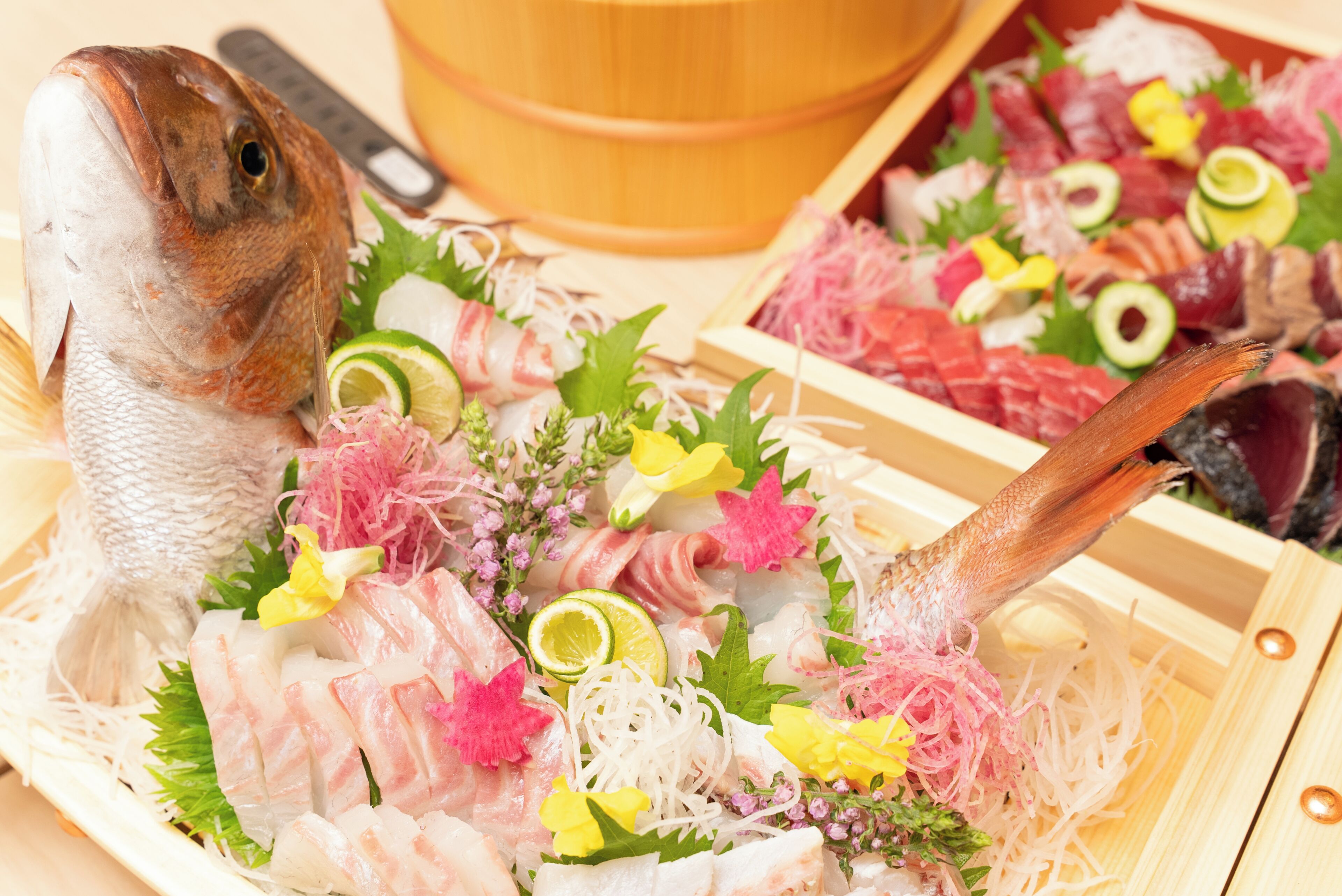 A variety of sushi and sashimi on a wooden tray with a fish head and tail, garnished with flowers and thinly sliced vegetables.