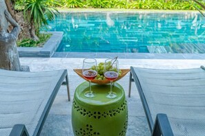 Ein Poolbereich mit zwei Liegestühlen, einem grünen Zierhocker und einem Korb voller Früchte und Getränke.