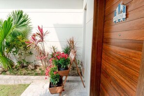 Eine Holztür mit einem 'WELCOME'-Schild, ein Blumentopf mit roten Blüten und ein kleiner Garten mit verschiedenen Pflanzen.