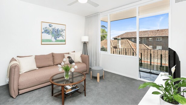 A modern living room with a beige sofa, a glass coffee table, and a view of a residential area through large windows.