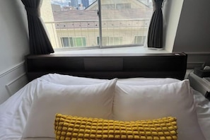 A bed with white bedding and a yellow accent pillow, a window with curtains, and a view of buildings outside. - Ikebukuro No`3 (Tokyo)