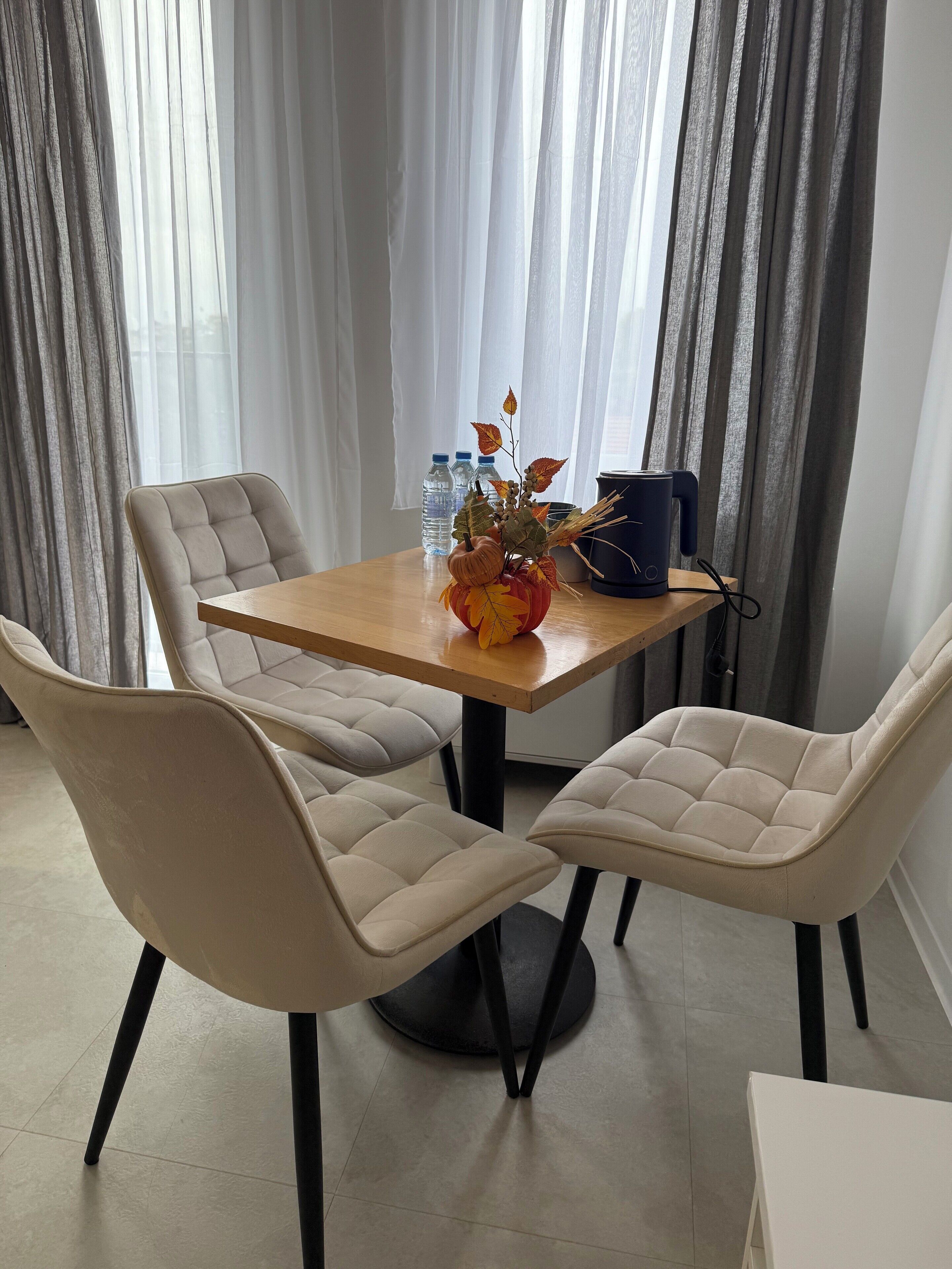 A dining area with a wooden table, four beige upholstered chairs, a vase with autumn leaves, and a blue coffee maker.
