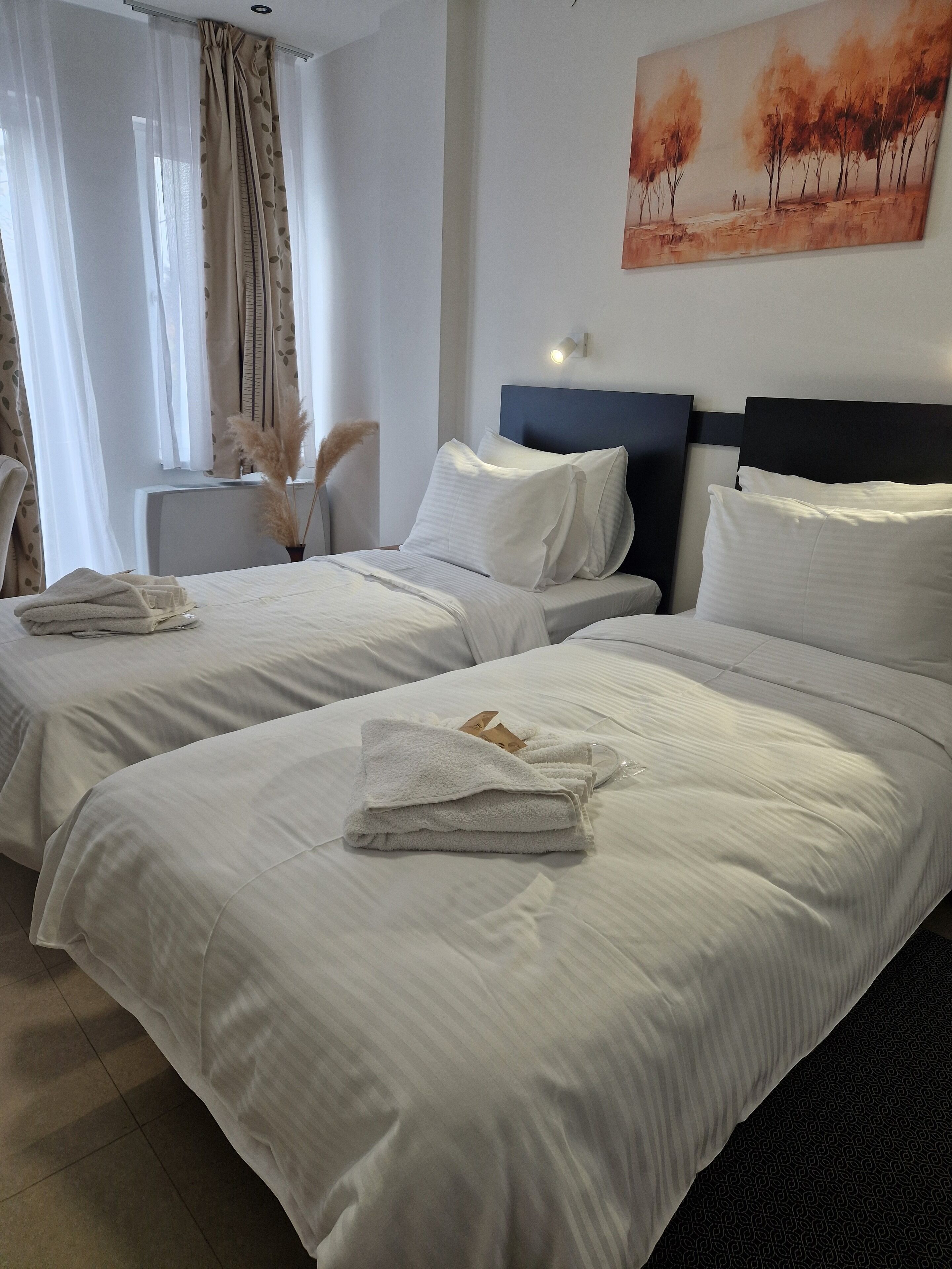A hotel room with two beds, white linens, a painting of trees, and a small side table with a vase of pampas grass.