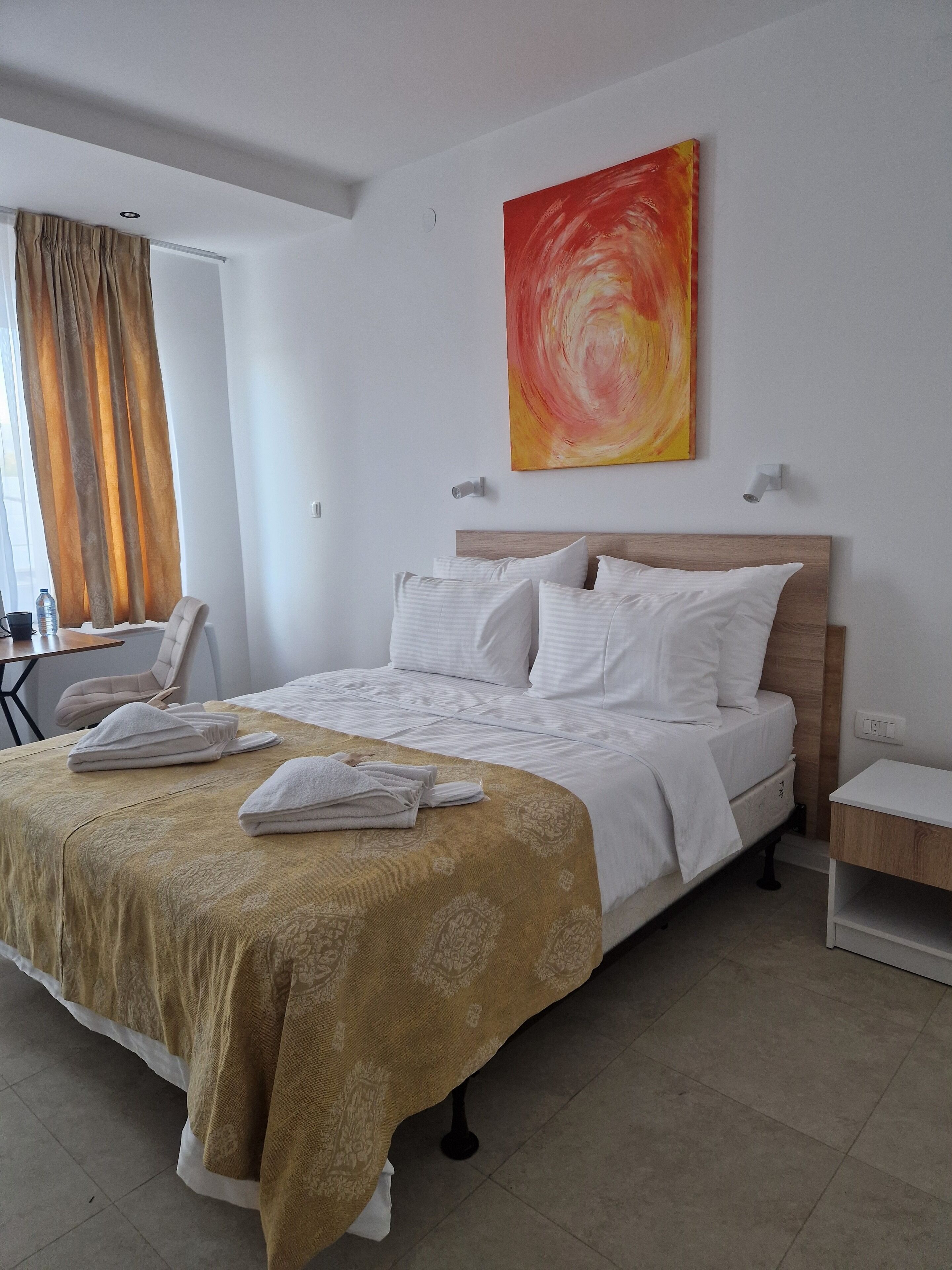 Een opgemaakt bed met witte lakens en een beige deken, een houten hoofdbord, en een groot abstract schilderij aan de muur.