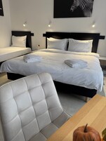 Habitación de hotel con dos camas, una butaca tapizada de blanco, una mesa de madera y una obra de arte con estampado de cebra en la pared.