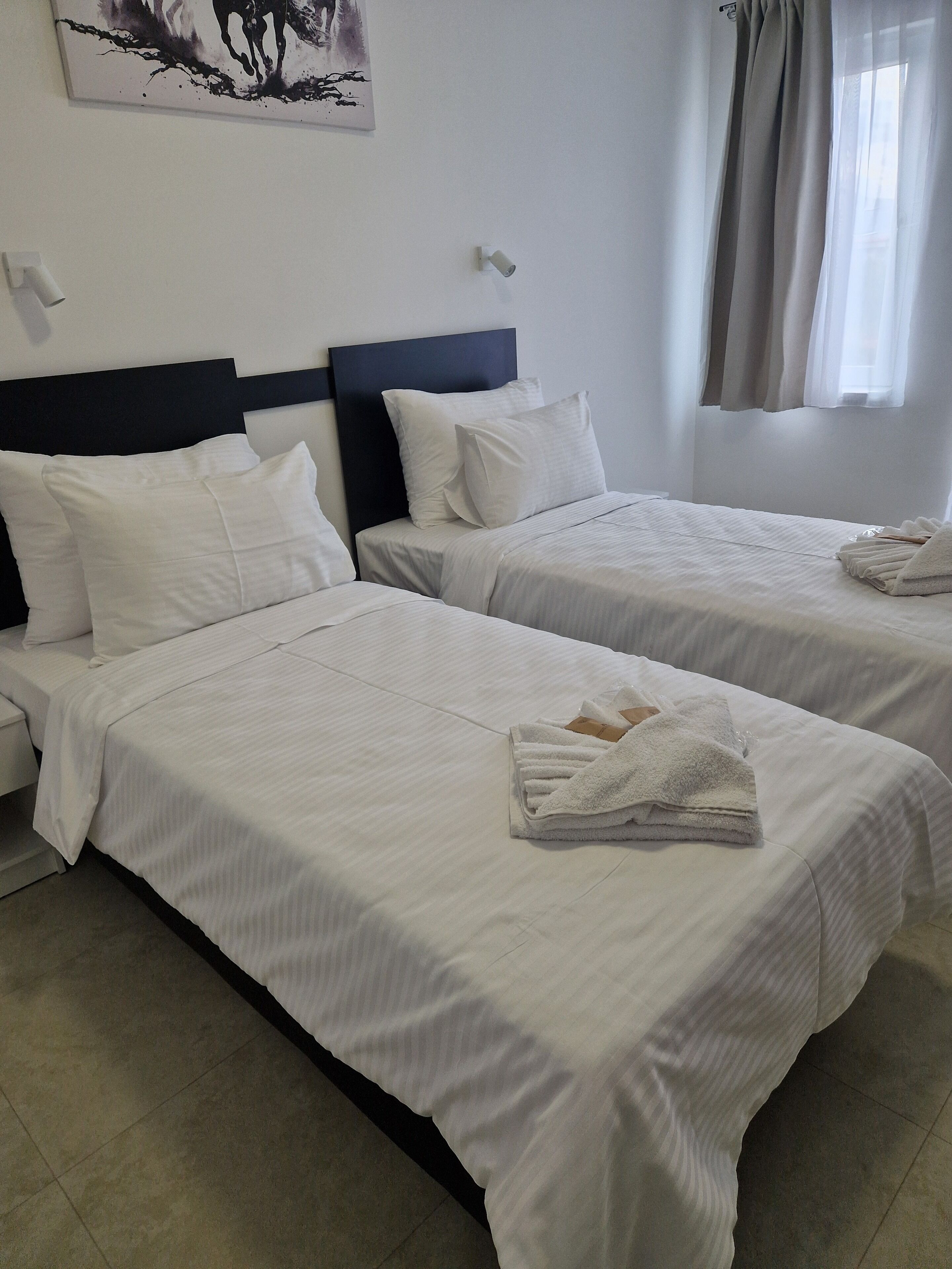 Une chambre d’hôtel avec deux lits, du linge de lit blanc, une tête de lit sombre, un tableau au mur et une fenêtre avec des rideaux.