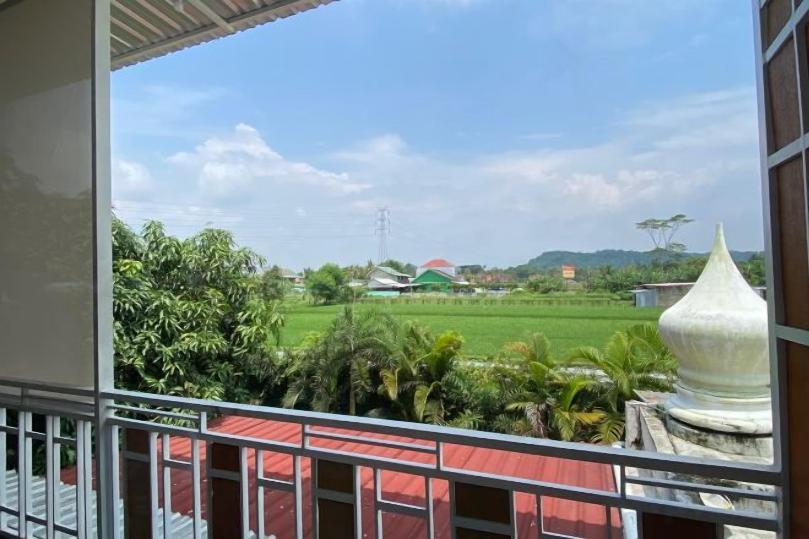 A view from a balcony with a red railing, overlooking a green field and a building with a green roof.