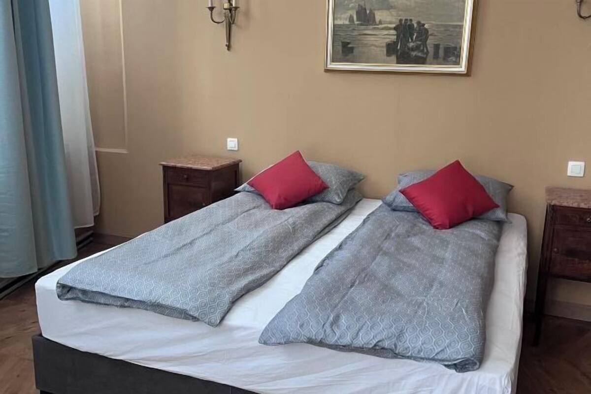 A double bed with grey and white bedding, two red pillows, and a painting on the wall.