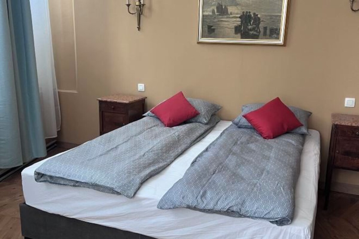A double bed with grey and white bedding, two red pillows, and a painting on the wall.