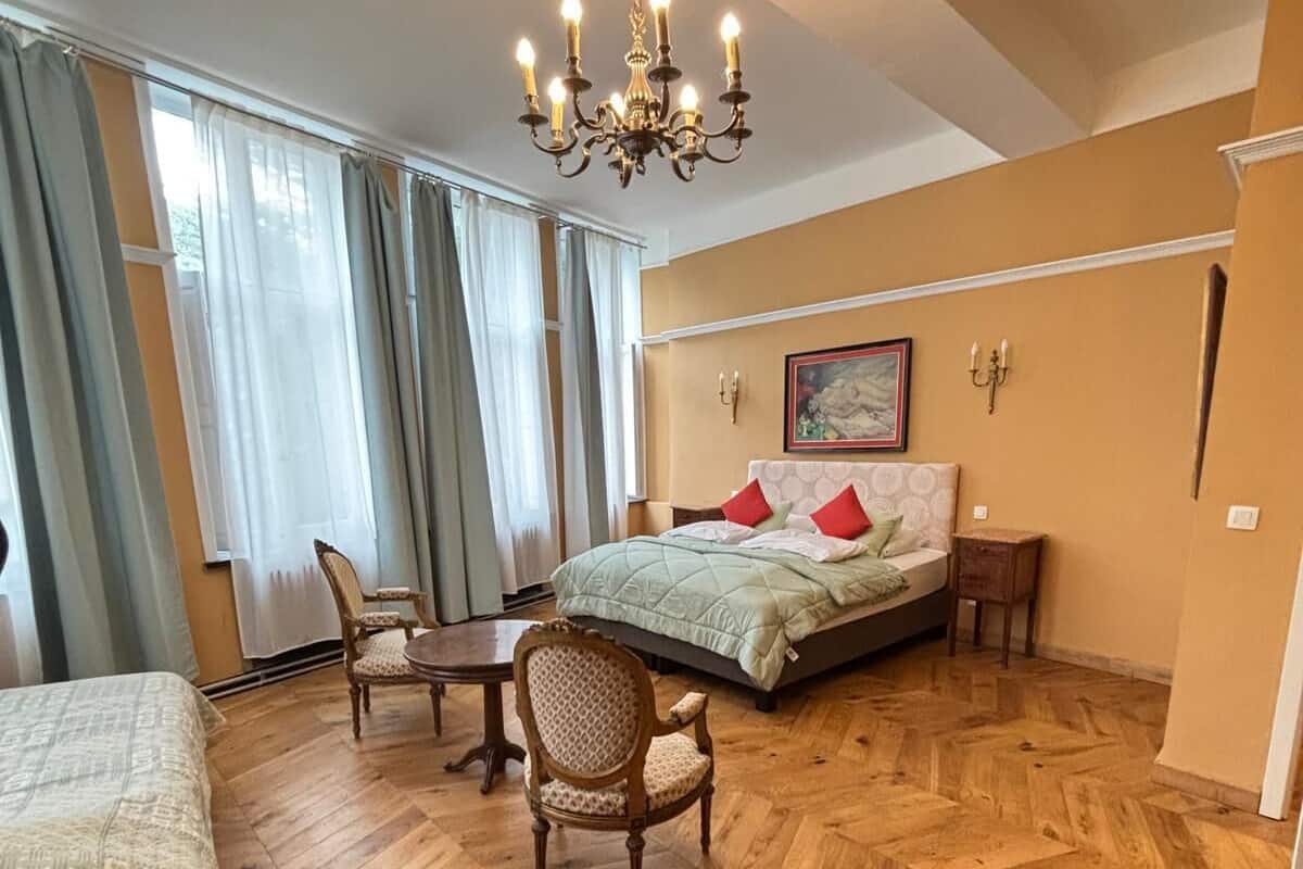 A bedroom with a large bed, a chair, a small table, a chandelier, and a painting on the wall.