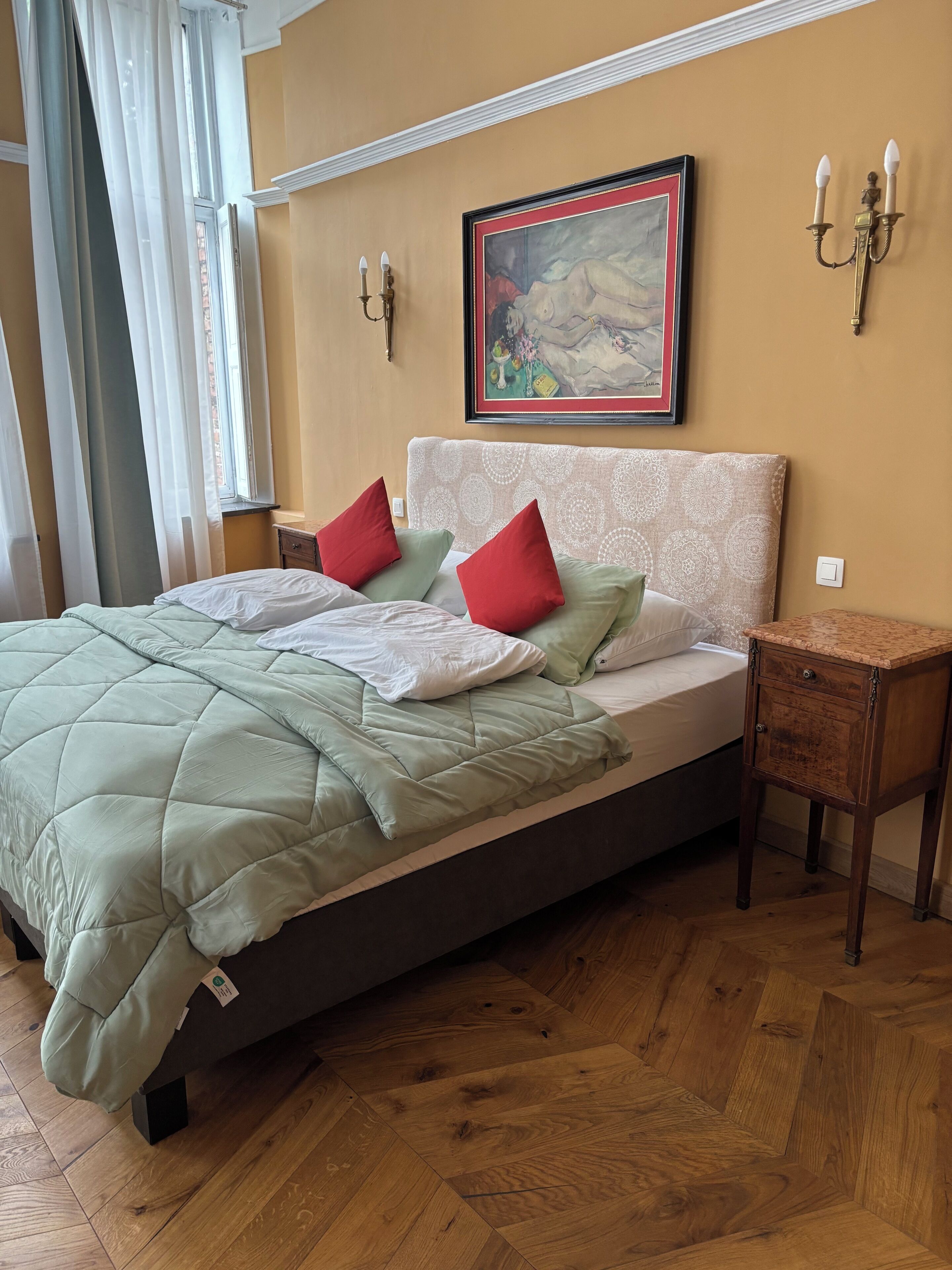 A bedroom with a bed, a nightstand, a wall-mounted lamp, and a painting.
