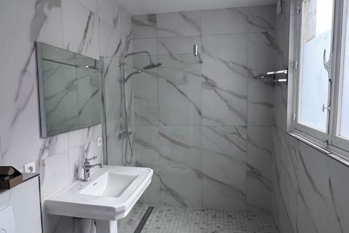 A bathroom with a marble wall, a sink, a shower, and a window.