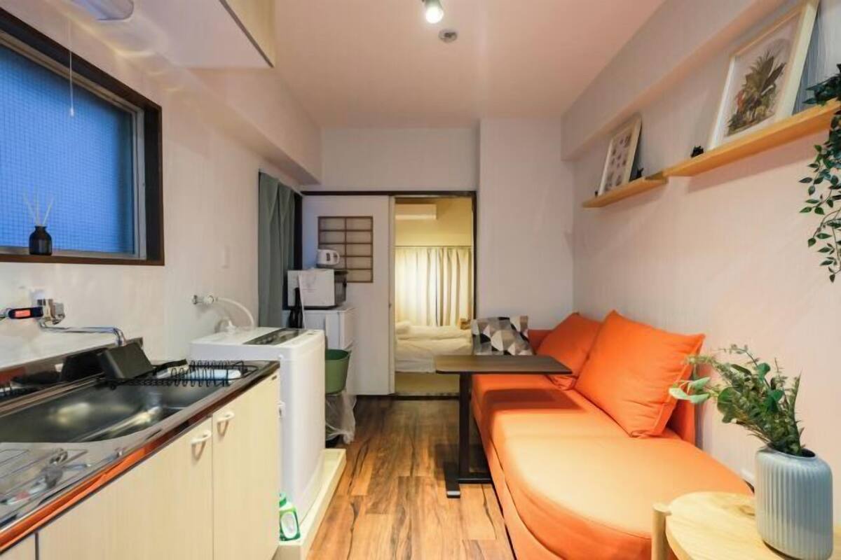 Un espacio habitable compacto con kitchenette, un sofá, una pequeña mesa de comedor y una ventana con persianas.