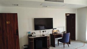 A hotel room with a wooden door, a flat-screen TV mounted on the wall, a desk with a chair, and a small trash bin. - Huadu Hotel (Kathmandu)