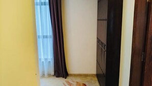 A room with a yellow wall, a window with white and dark curtains, and a wooden door. - Huadu Hotel (Kathmandu)