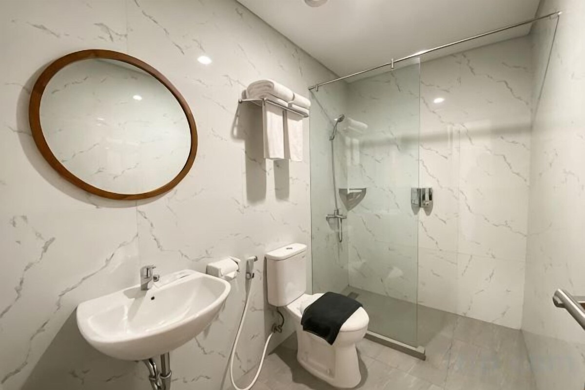 A modern bathroom with a marble wall, a round mirror, a sink, a toilet, and a glass shower enclosure.