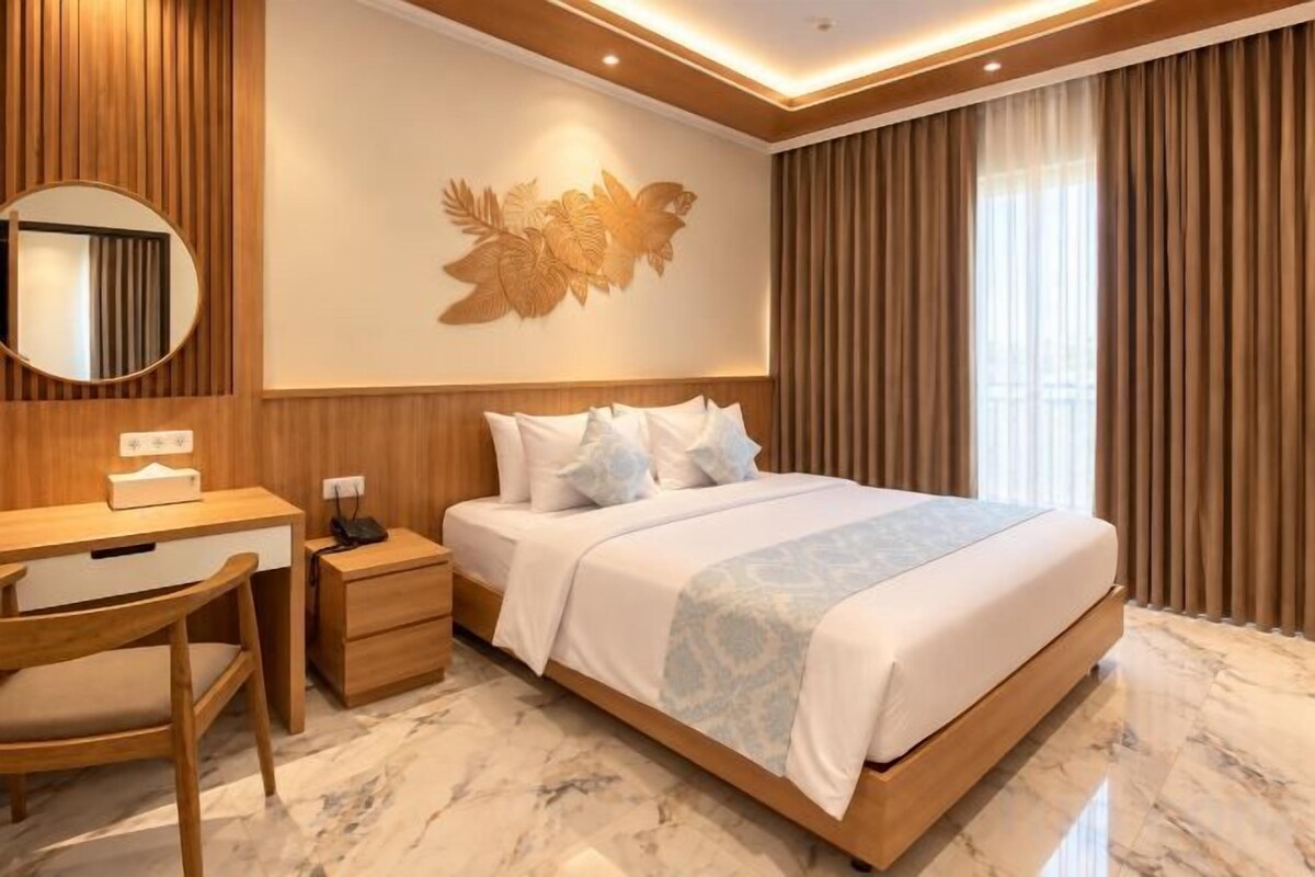 A hotel room with a bed, a desk, a chair, a nightstand, and a wall-mounted mirror.